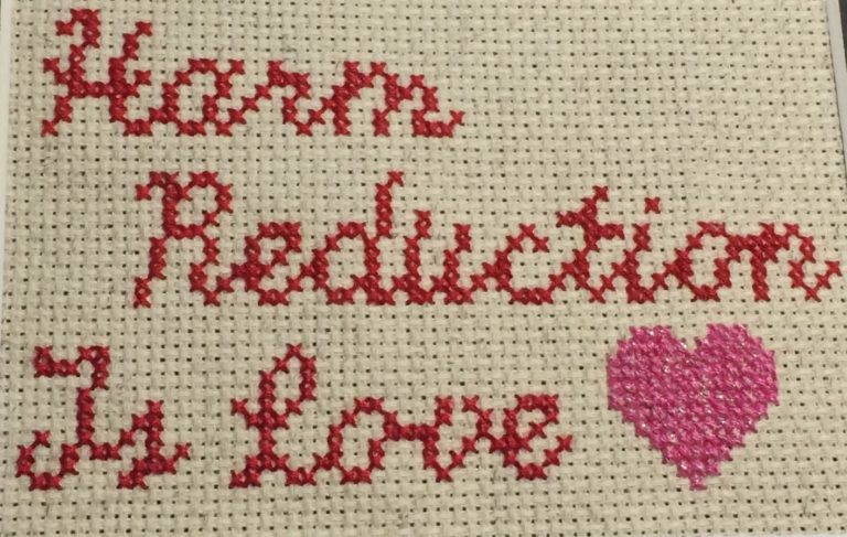 Cross-stiched red handwriting that says "harm reduction is love" followed by a pink heart