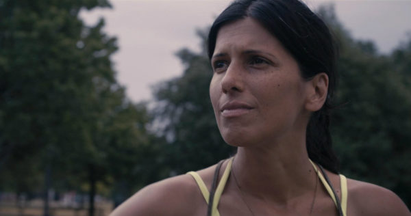 Attiya Khan looks into the distance, with blurry everygreen trees in the background. She squints against the light. Attiya Khan is the co-director of the film and her story is the basis of the documentary