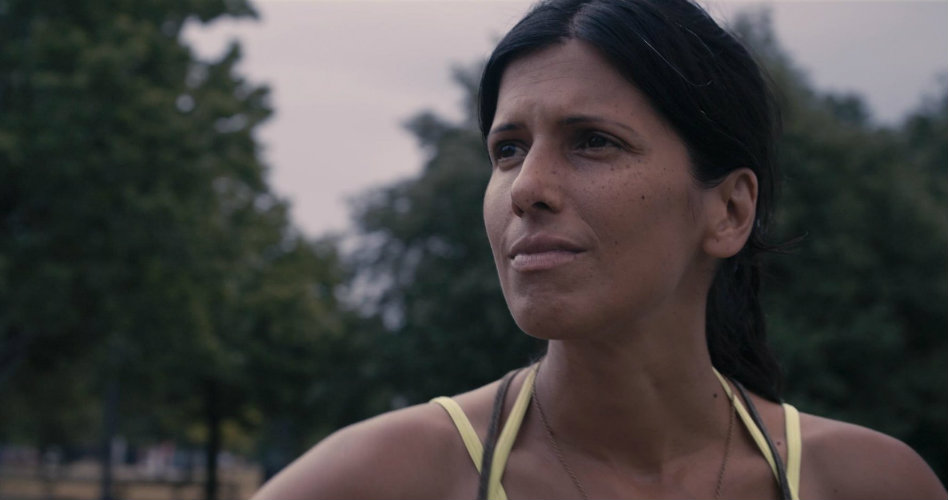 Attiya Khan looks into the distance, with blurry everygreen trees in the background. She squints against the light. Attiya Khan is the co-director of the film and her story is the basis of the documentary