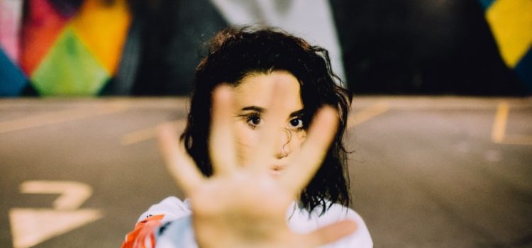 Feeling through rejection Image of a person with their hand extended out in front of them with their fingers spread out as if to signify "stop" or "no." Behind the person is in parking lot that has colourful walls