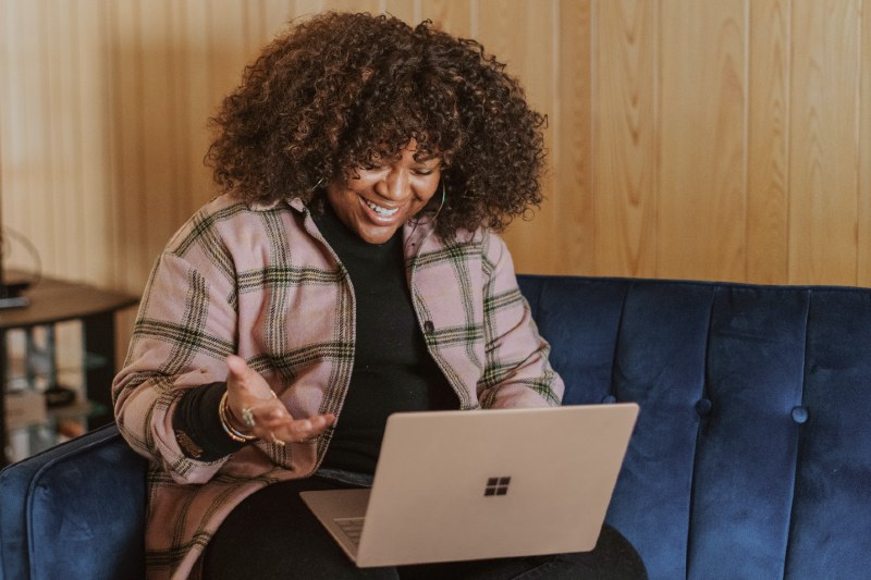 A person sitting on a blue couch smiles at their open laptop screen