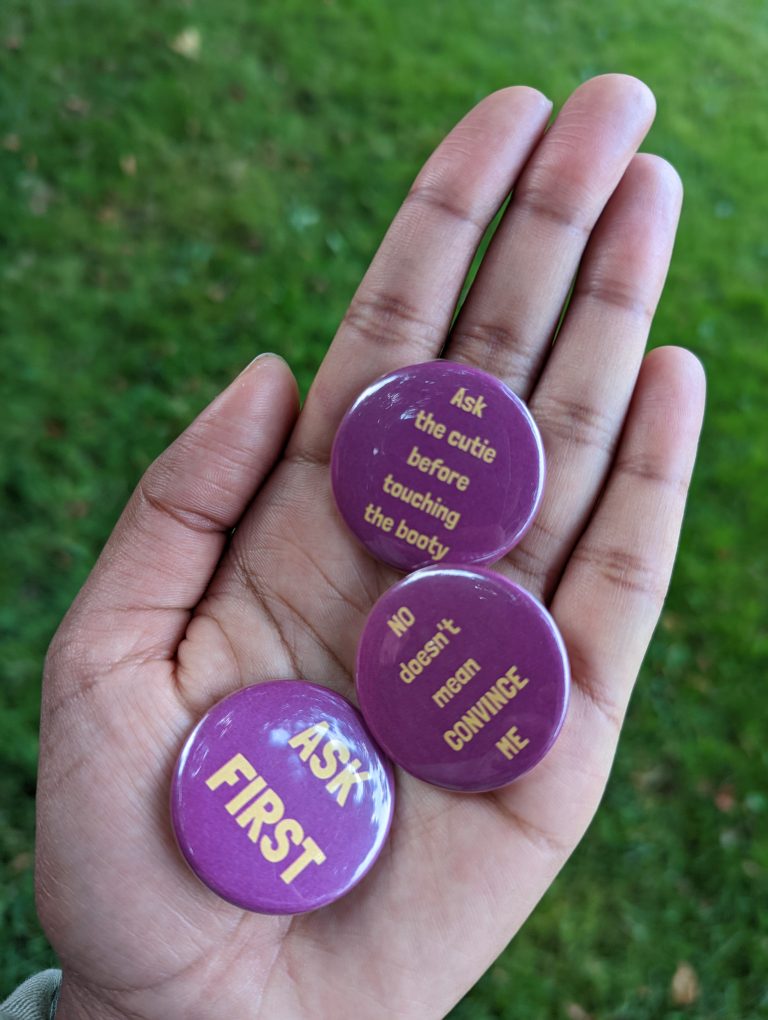 An open hand holding three buttons with three different phrases. "ask first," "no does not mean convince me," and "ask the cutie before touching the booty."
